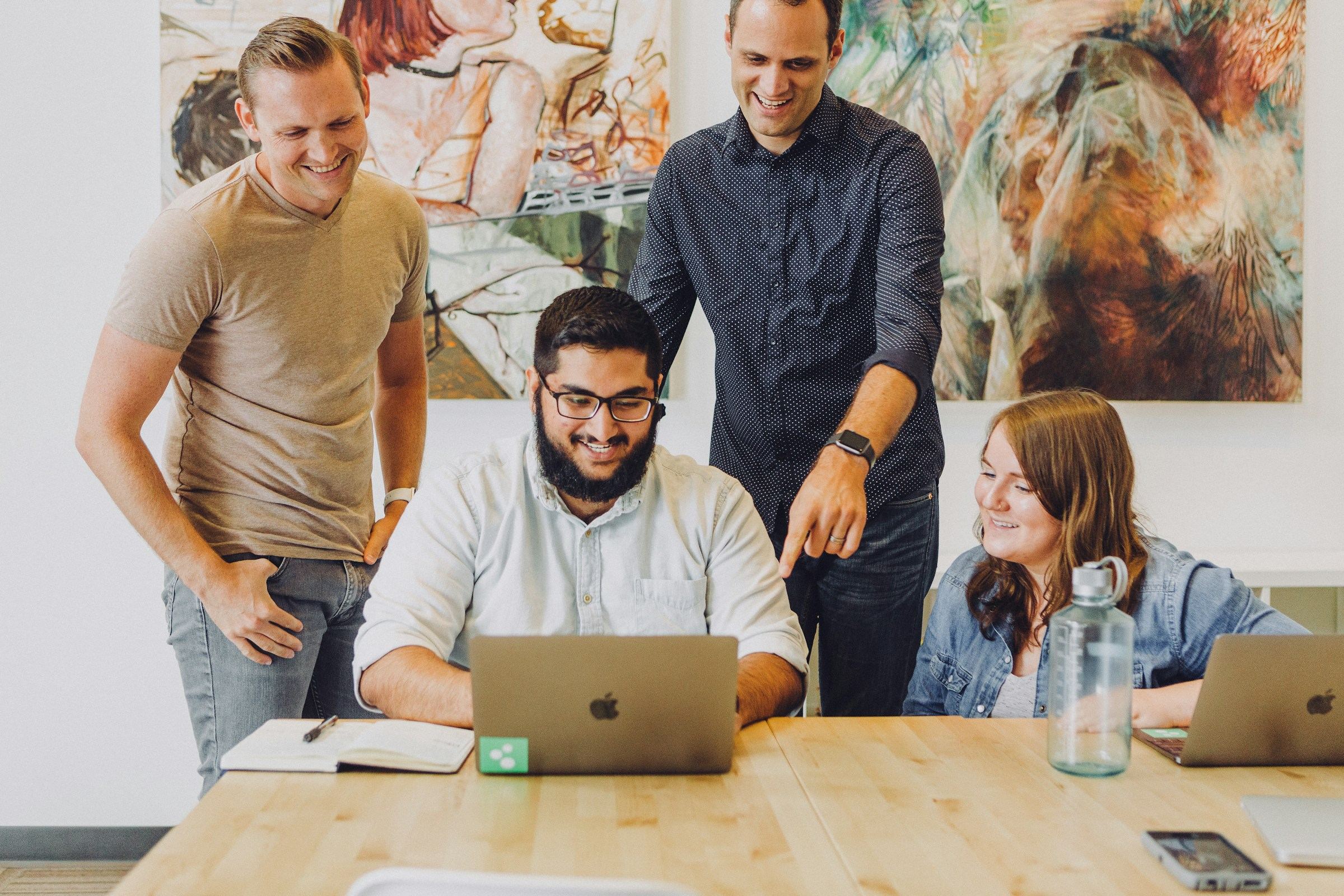 Four smiling colleagues reviewing a laptop together at a table.